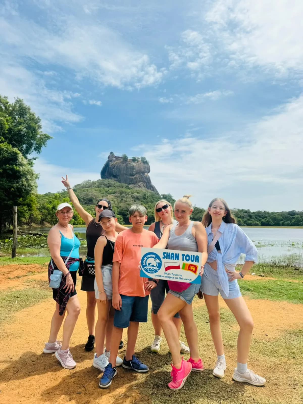 A group holding the Shan Tours board with Sigiriya rock behind them